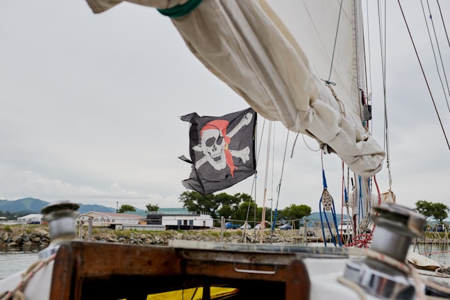 A pirate flag featuring a skull and crossbones design with a red bandana is prominently displayed on a sailboat. The background includes a marina with several buildings, trees, and distant hills under a cloudy sky. Part of the boat's fixtures, such as ropes and a winch, are visible in the foreground.