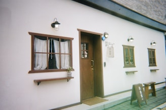 A quaint building facade with a wooden door and two windows framed with dark wood. The windows have sheer curtains, and a small plant decorates one of the windowsills. Exterior lights adorn the walls above. A simple sign with text in a non-Latin script hangs beside the door, and a wooden sandwich board stands on the pavement.