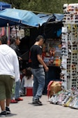 A bustling street market scene features a group of people gathered around a stall selling an assortment of sunglasses and bottled drinks. The background includes colorful umbrellas providing shade, and various products neatly arranged for display. The atmosphere suggests a busy and lively environment typical of market areas.