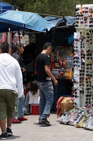 A bustling street market scene features a group of people gathered around a stall selling an assortment of sunglasses and bottled drinks. The background includes colorful umbrellas providing shade, and various products neatly arranged for display. The atmosphere suggests a busy and lively environment typical of market areas.