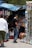 A bustling street market scene features a group of people gathered around a stall selling an assortment of sunglasses and bottled drinks. The background includes colorful umbrellas providing shade, and various products neatly arranged for display. The atmosphere suggests a busy and lively environment typical of market areas.
