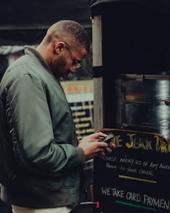 A man in a green jacket and glasses stands in front of a food stall, typing on his phone. The stall's sign mentions card payments and instructions for any allergies prior to ordering. The setting appears urban and casual.