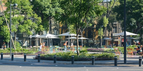 A vibrant urban park setting with colorful chairs and tables scattered under large umbrellas. Trees surround the area, providing shade, and a few people are enjoying the outdoor space. Buildings with classic architecture are visible in the background.
