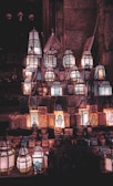 A display of intricately designed lanterns illuminated with a warm glow, showcasing various geometric patterns and shapes. The lanterns are arranged on a step-like surface against a textured background, possibly stone walls. The ambient light enhances the ornate detailing, creating a mystical atmosphere.