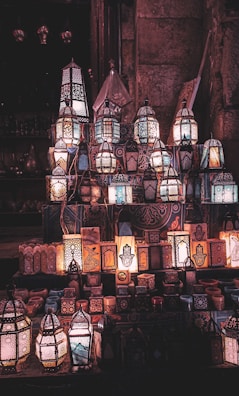 A display of intricately designed lanterns illuminated with a warm glow, showcasing various geometric patterns and shapes. The lanterns are arranged on a step-like surface against a textured background, possibly stone walls. The ambient light enhances the ornate detailing, creating a mystical atmosphere.