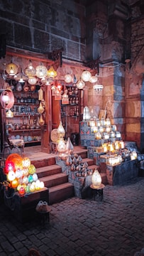 A vibrant market stall featuring a variety of traditional lamps, displayed both hanging and on tables. The stall is located within a rustic building with stone walls, creating an enchanting atmosphere. The lamps are illuminated, showcasing intricate designs and casting a warm glow.