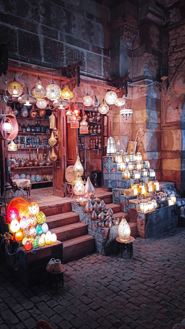 A vibrant market stall featuring a variety of traditional lamps, displayed both hanging and on tables. The stall is located within a rustic building with stone walls, creating an enchanting atmosphere. The lamps are illuminated, showcasing intricate designs and casting a warm glow.