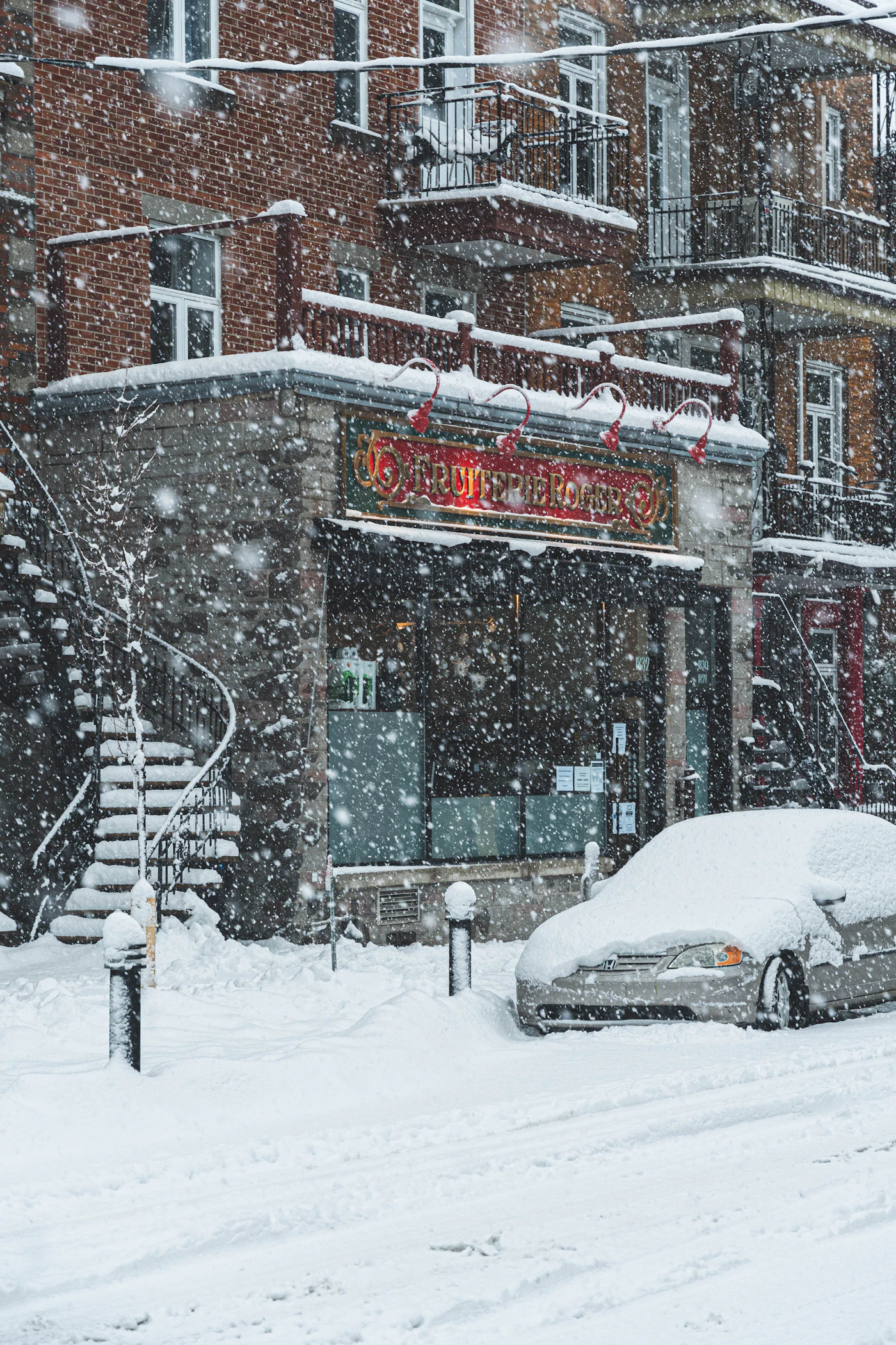 A corner depanneur and brick triplex with an iron exterior staircase during heavy snowfall on a Montreal street, a parked car buried in snow
