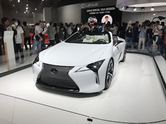 A sleek white Lexus convertible is on display in a showroom. It is surrounded by a large crowd of people, some of whom are taking photos. The car features a modern, aerodynamic design with sharp lines and a prominent front grille. Overhead lighting casts reflections on the car’s glossy surface.
