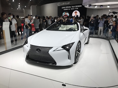 A sleek white Lexus convertible is on display in a showroom. It is surrounded by a large crowd of people, some of whom are taking photos. The car features a modern, aerodynamic design with sharp lines and a prominent front grille. Overhead lighting casts reflections on the car’s glossy surface.