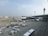 An airport scene with several airplanes positioned at the terminal gates, with a prominent airplane from ANA in blue and white livery. Various airport vehicles such as luggage carriers and shuttle buses are scattered around the tarmac. A large control tower is visible in the background, and the sky is clear with some clouds.