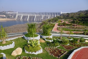 A large concrete dam is situated in the background, with water flowing beneath it. The foreground features a beautifully landscaped garden with various green plants and shrubs. Curved, red pathways wind through the garden, bordered by manicured hedges and small trees. The sky is clear and blue, indicating a sunny day.
