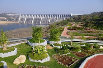 A large concrete dam is situated in the background, with water flowing beneath it. The foreground features a beautifully landscaped garden with various green plants and shrubs. Curved, red pathways wind through the garden, bordered by manicured hedges and small trees. The sky is clear and blue, indicating a sunny day.