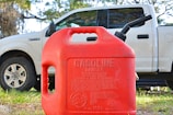 A red plastic gasoline container with a warning label is prominently placed in the foreground. Behind it, a white pickup truck is partially visible, parked on a grassy area with trees in the background.