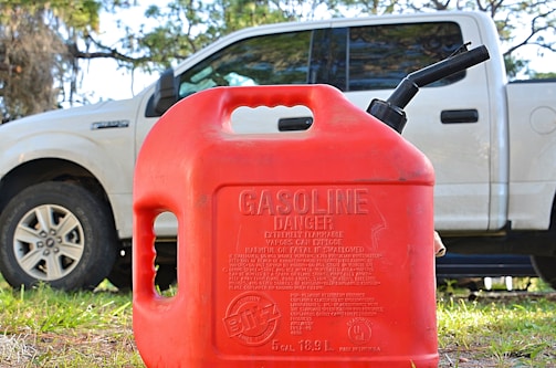 A red plastic gasoline container with a warning label is prominently placed in the foreground. Behind it, a white pickup truck is partially visible, parked on a grassy area with trees in the background.