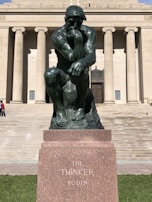 A bronze statue of a contemplative man sits on a stone base. The background features a neoclassical building with large columns and a set of stairs leading up to the entrance.