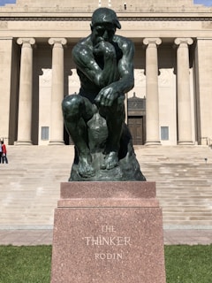 A bronze statue of a contemplative man sits on a stone base. The background features a neoclassical building with large columns and a set of stairs leading up to the entrance.