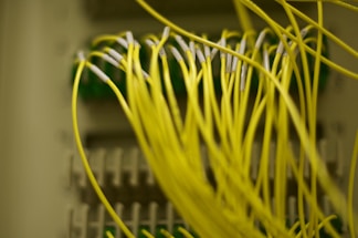 A close-up image of numerous yellow fiber optic cables connected to ports on a network panel. The cables are prominently in focus, with some parts of the panel appearing blurred in the background.