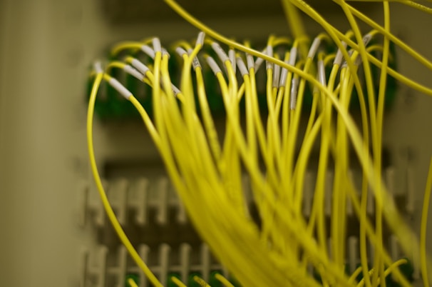 A close-up image of numerous yellow fiber optic cables connected to ports on a network panel. The cables are prominently in focus, with some parts of the panel appearing blurred in the background.