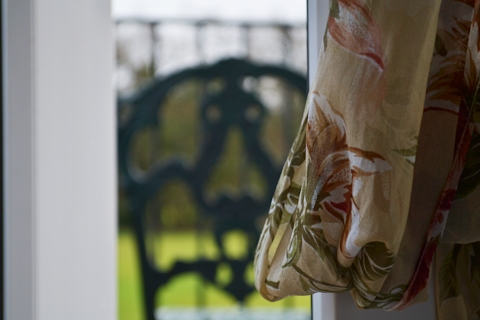 A delicate floral-patterned curtain is partially drawn to the side, allowing soft light to filter through. The window reveals a green blurred outdoor scenery. The curtain&rsquo;s fabric features earthy tones and intricate floral designs, creating a warm and cozy atmosphere.