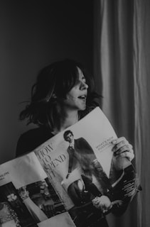 A person holds a magazine and appears to be engaged with it. The magazine cover features a headline 'How to Spend It' and images related to fashion and lifestyle, including a stylishly dressed individual and a cat. The setting is indoors with a curtain visible in the background.