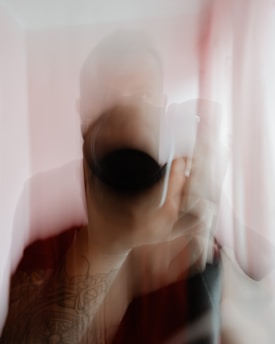 A blurred image showing a person holding a camera with visible tattooed arms. The photograph has a soft focus, creating an abstract effect with subtle movement.