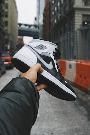 A hand holds a black and white sneaker, prominently featuring the Air Jordan logo, against an urban street background with buildings and a bridge visible in the distance. The foreground is in focus while the background is slightly blurred, adding depth to the scene.