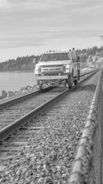 A utility truck is positioned on a railway track, surrounded by rocky terrain and bordered by a metal fence. The background features a body of water and a tree line, under a cloudy sky.