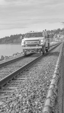 A utility truck is positioned on a railway track, surrounded by rocky terrain and bordered by a metal fence. The background features a body of water and a tree line, under a cloudy sky.