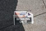 A newspaper lies on a concrete sidewalk with a headline reading 'WORKING ON IT'. The newspaper appears partially crumpled with its pages curling at the corner. It features a mix of photographs and text, including an image of a person in the top section. Sunlight casts a shadow across part of the newspaper and the sidewalk, adding to the scene's contrast.