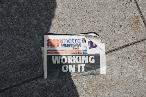 A newspaper lies on a concrete sidewalk with a headline reading 'WORKING ON IT'. The newspaper appears partially crumpled with its pages curling at the corner. It features a mix of photographs and text, including an image of a person in the top section. Sunlight casts a shadow across part of the newspaper and the sidewalk, adding to the scene's contrast.