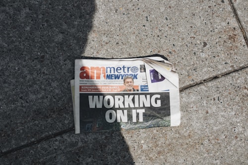 A newspaper lies on a concrete sidewalk with a headline reading 'WORKING ON IT'. The newspaper appears partially crumpled with its pages curling at the corner. It features a mix of photographs and text, including an image of a person in the top section. Sunlight casts a shadow across part of the newspaper and the sidewalk, adding to the scene's contrast.
