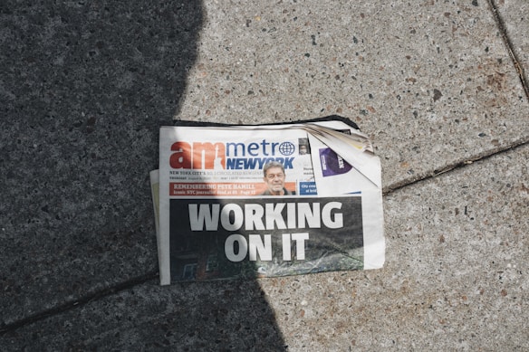 A newspaper lies on a concrete sidewalk with a headline reading 'WORKING ON IT'. The newspaper appears partially crumpled with its pages curling at the corner. It features a mix of photographs and text, including an image of a person in the top section. Sunlight casts a shadow across part of the newspaper and the sidewalk, adding to the scene's contrast.