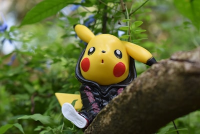 A toy figure of Pikachu wearing a black hoodie is perched on a tree branch amidst lush green foliage. The character's bright yellow color and red cheeks stand out against the natural background.