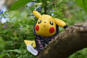 A toy figure of Pikachu wearing a black hoodie is perched on a tree branch amidst lush green foliage. The character's bright yellow color and red cheeks stand out against the natural background.