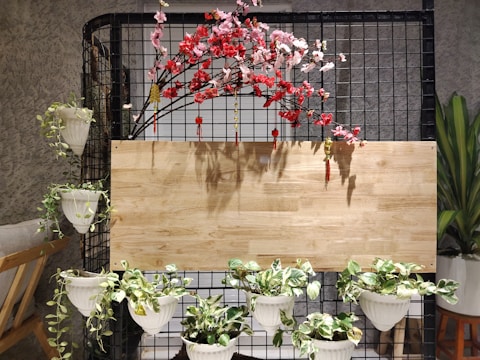 A decorative setup featuring a black grid structure adorned with artificial cherry blossom branches in pink and red. Small decorative tassels hang from the branches. Several lush green plants are placed in white planters hanging from the structure and positioned at the bottom. A wooden board serves as a backdrop, adding a rustic touch to the arrangement. A chair with a cushion sits on the left side, and a tall plant is visible on the right.