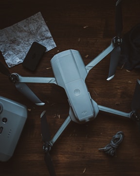 A drone with four propellers is placed on a dark wooden surface. A rope is coiled nearby, along with a crumpled map and a remote control device.