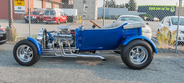 A vintage blue hot rod with shiny chrome detailing and exposed engine parts is parked outdoors on a paved surface, alongside a chain-link fence. In the background, there are a fire rescue vehicle and a few other cars, as well as buildings with signage.