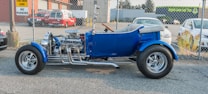 A vintage blue hot rod with shiny chrome detailing and exposed engine parts is parked outdoors on a paved surface, alongside a chain-link fence. In the background, there are a fire rescue vehicle and a few other cars, as well as buildings with signage.