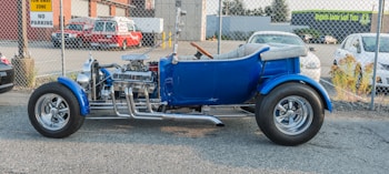 A vintage blue hot rod with shiny chrome detailing and exposed engine parts is parked outdoors on a paved surface, alongside a chain-link fence. In the background, there are a fire rescue vehicle and a few other cars, as well as buildings with signage.