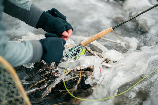 Angler in dark fold-over fleece gloves with bare fingertips rigging a fly rod over ice-glazed rocks, green fly line trailing across the ice