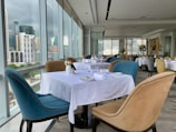 Elegant dining area with large windows offering a cityscape view. Tables are set with white tablecloths, neatly arranged plates, glasses, and cutlery. Upholstered chairs in blue and beige surround the tables. Flower vases with floral arrangements decorate the space, creating a sophisticated atmosphere.