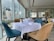 Elegant dining area with large windows offering a cityscape view. Tables are set with white tablecloths, neatly arranged plates, glasses, and cutlery. Upholstered chairs in blue and beige surround the tables. Flower vases with floral arrangements decorate the space, creating a sophisticated atmosphere.