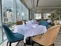 Elegant dining area with large windows offering a cityscape view. Tables are set with white tablecloths, neatly arranged plates, glasses, and cutlery. Upholstered chairs in blue and beige surround the tables. Flower vases with floral arrangements decorate the space, creating a sophisticated atmosphere.