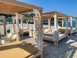 A beach setting with several wooden cabanas featuring white curtains and lounge chairs. The shore is covered with pebbles, and the sea is visible in the background under a clear blue sky. There are some people enjoying the beach and a nearby structure painted in green and red.