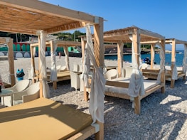A beach setting with several wooden cabanas featuring white curtains and lounge chairs. The shore is covered with pebbles, and the sea is visible in the background under a clear blue sky. There are some people enjoying the beach and a nearby structure painted in green and red.