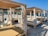 A beach setting with several wooden cabanas featuring white curtains and lounge chairs. The shore is covered with pebbles, and the sea is visible in the background under a clear blue sky. There are some people enjoying the beach and a nearby structure painted in green and red.