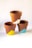 Three terracotta flower pots are stacked in a pyramid formation against a plain white background. Each pot is partially painted with a triangular pattern in different colors: one with white, one with yellow, and one with blue.