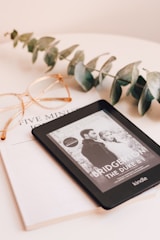 A Kindle device displaying a book titled 'Bridgerton: The Duke & I' rests on top of a closed book titled 'Five Min'. Beside the Kindle, there is a pair of eyeglasses with a gold frame and a sprig of eucalyptus leaves, all placed on a pale beige surface.
