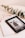 A Kindle device displaying a book titled 'Bridgerton: The Duke & I' rests on top of a closed book titled 'Five Min'. Beside the Kindle, there is a pair of eyeglasses with a gold frame and a sprig of eucalyptus leaves, all placed on a pale beige surface.